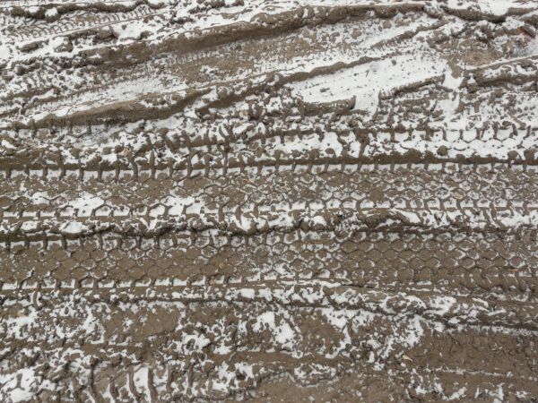 Dirt road texture in dark brown tone with tire marks and thin layer of snow across surface.